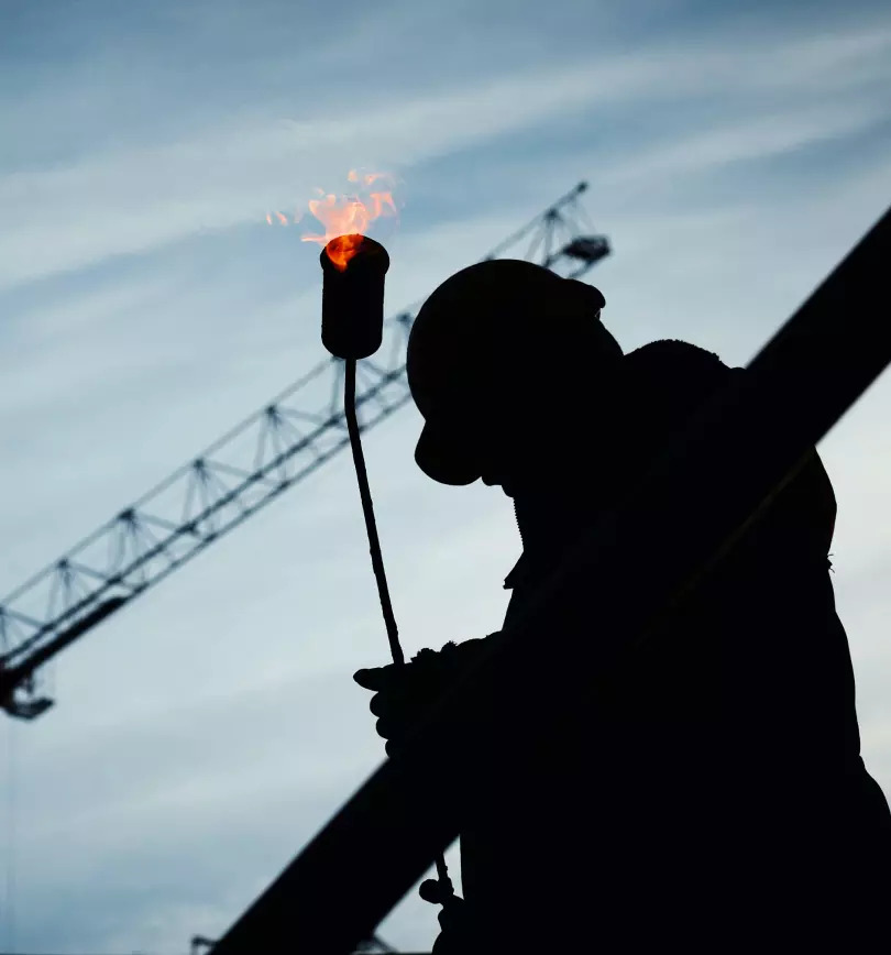 A silhouette of a worker in a hard hat holding a torch with a crane in the background