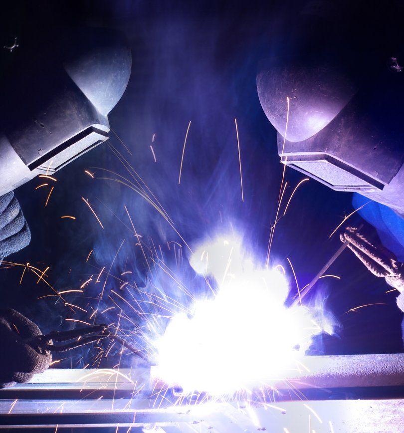 Two welders in face shields working together with a bright flame in between them